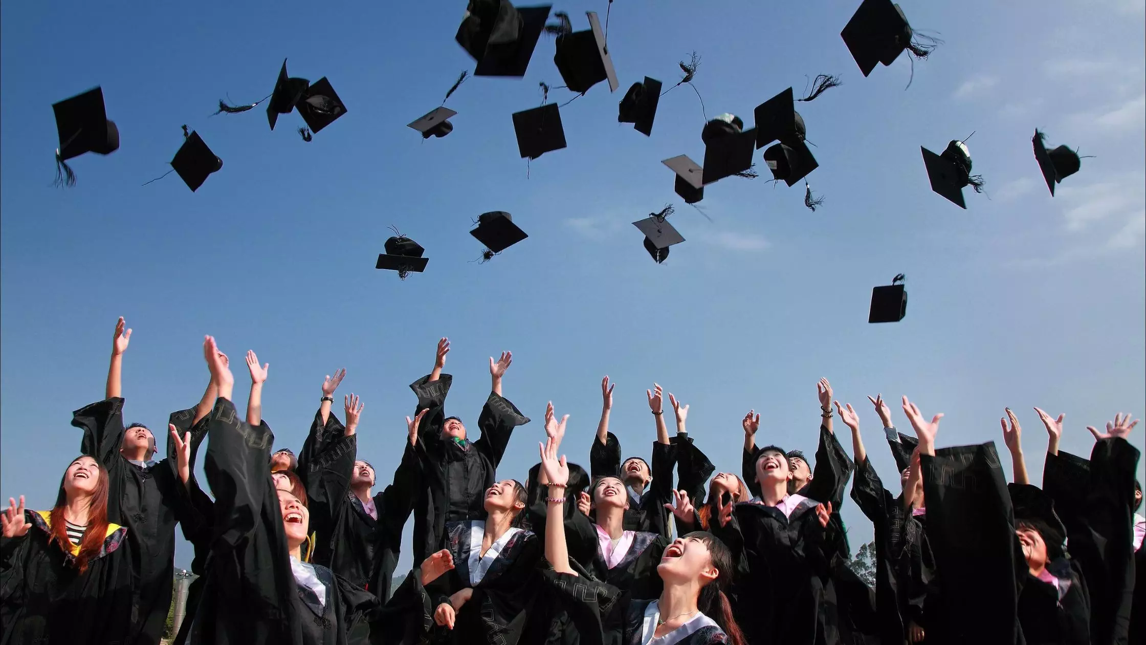 A large group of graduates celebrating in caps and gowns, throwing their black graduation caps high in the air.